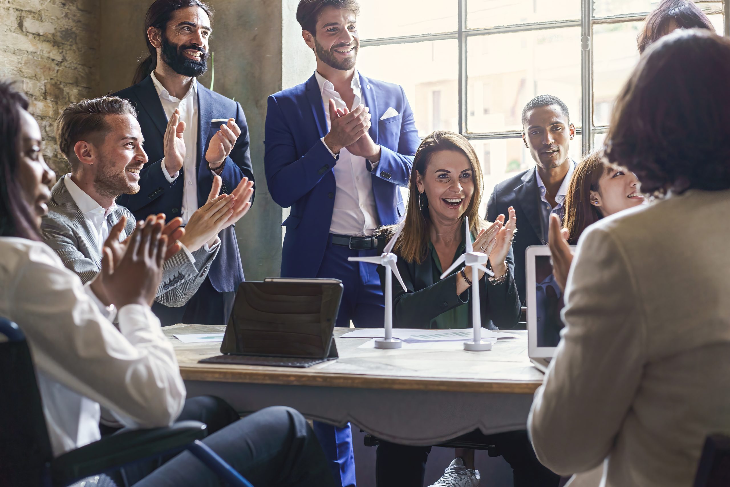 Group of multiethnic coworkers applauding the success of a sustainability project about renewable energy and green power – teamwork clapping hands sitting around the table of the meeting room
