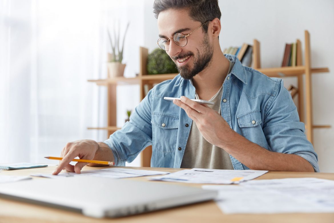 Portrait of cheerful bearded man has voice call, speaks with friend and checks documents, has happy expression. Hardworking male checks finances, does accounting, sits at desk with papers. Job concept
